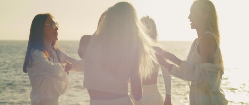 Four women enjoy a fun conversation on the beach at sunset, capturing the moment in soft golden light and ocean breeze.