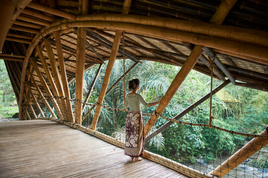Asia, Indonesia, Bali, Ubud, Mature Asian female tourist standing on bamboo bridge crossing small river, wearing asian style clothing