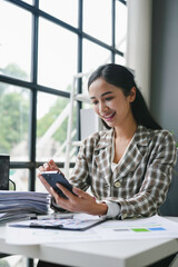 Asian businesswoman is using a smartphone and smiling while working at a desk in a modern office