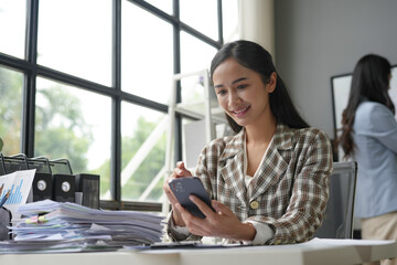 Young manager is smiling while using her smartphone in a busy office with her colleague working in...