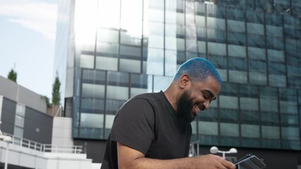 Smiling african american young man in casual outfit blue hair bursts out laughing in a video call with teammate about successful financial project with modern buildings environment.MOV - Powered by Adobe