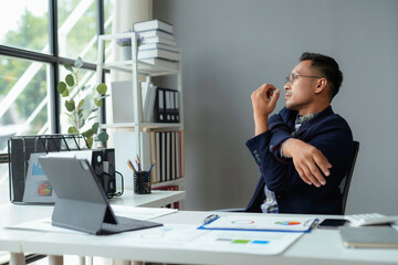 Businessman stretching arms at office desk with documents and digital tablet in bright office space