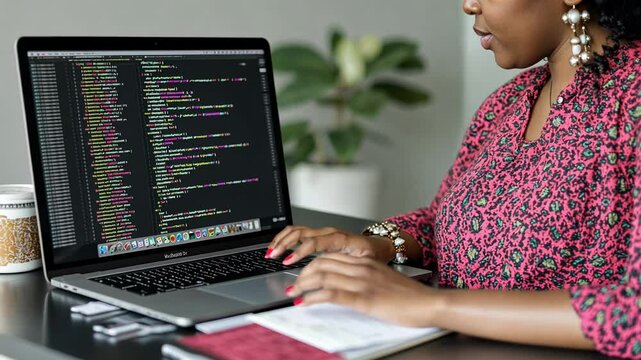 A woman is focused on writing code on her laptop at a modern desk. The workspace features indoor plants that add a fresh ambiance to the environment.