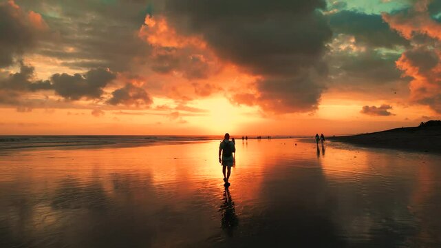 Aerial view of people walking on a Bali beach during a stunning sunset with dramatic clouds reflected in the water.