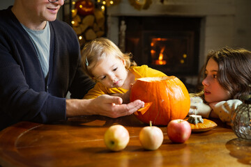Fototapeta premium Father his toddler and daughter carving pumpkin jack o lantern, Halloween celebration at home. Fun craft and decoration at home. Happy children and their dad preparing for the holiday, decorating the