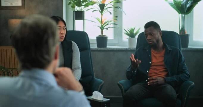 Multiethnic young family sit on chairs, discuss relationship with psychologist on counseling session. Back view of therapist listening to couple, providing couples therapy. Psychology and sexology.