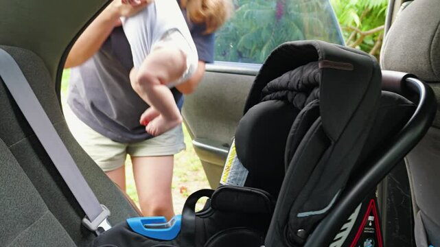 A woman wearing glasses picks up an infant from an infant car seat. The car seat is equipped with seat belts. The scene takes place in back seat of car. A mum takes her son out of car after a journey