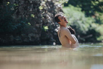 Fototapeta premium A delighted father holds his young daughter while laughing in a calm outdoor lake, surrounded by nature. The image captures happiness and the joy of family moments in a natural setting.