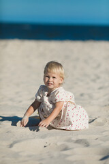 2 years old girl in white clothes sits on the white sand on the beach in summer