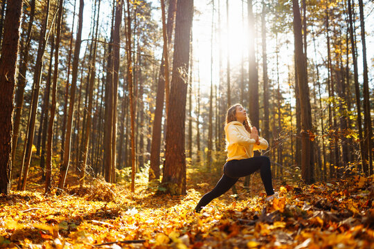 A serene woman practices yoga meditation in a sunlit forest surrounded by autumn leaves. Lifestyle and Meditation concept.