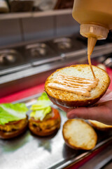 A close-up of a hand applying sauce to an open hamburger bun over a metal surface with prepared burger ingredients. The scene captures the process of assembling a delicious homemade burger