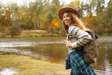 A young traveler woman exploring a serene forest path in autumn, wearing a cozy sweater and wide-brimmed hat with a backpack. Travel, nature concept.