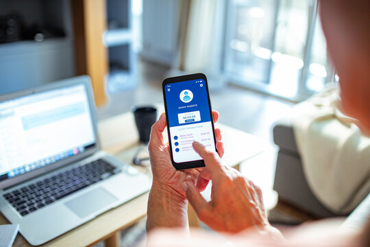 Senior woman checking bank account on smartphone with laptop in the background at home