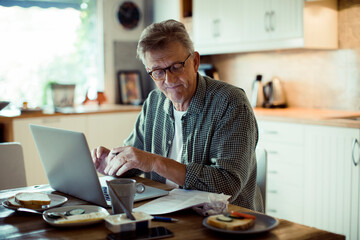 Stressed mature man using laptop on kitchen table during breakfast