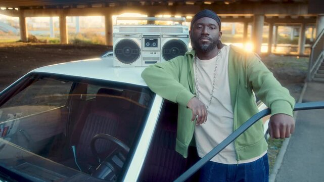 African American man in casualwear and durag leaning on white car with retro boombox on roof and looking confidently into camera under road bridge with warm sunshine in background. Video portrait
