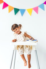 cute smiling girl 2 years old sitting in high chair on white background with colorful flags holding piece of birthday cake, makes a face, birthday concept, vertical photo
