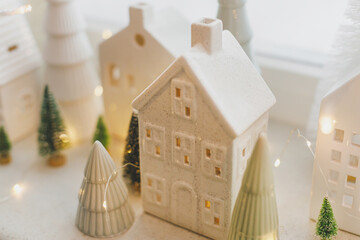 Merry Christmas! Stylish Christmas miniature holiday village with white houses, trees and golden lights bokeh on snowy window. Atmospheric magical scene. Christmas holiday banner.