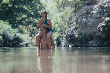 A joyful couple having fun in a tranquil river environment. The woman rides on the man's shoulders as they splash in the water, surrounded by natural beauty.