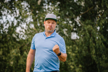 A man in a blue polo shirt makes a celebratory fist pump while making a playful face outdoors, with...