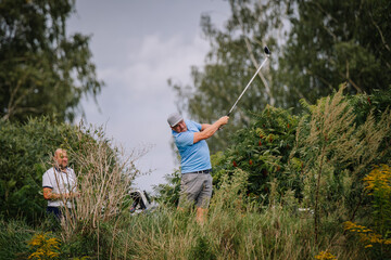 A golfer swings his club in a lush outdoor setting, surrounded by greenery, while another man observes in the background.