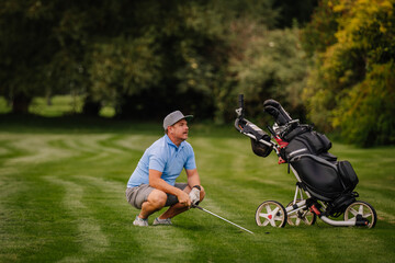 A golfer crouches on the fairway, analyzing his shot while holding a club. A golf cart with bags...
