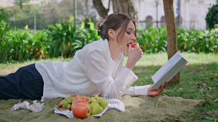 Reading girl lying blanket at green grass park eating apple closeup. Happy woman
