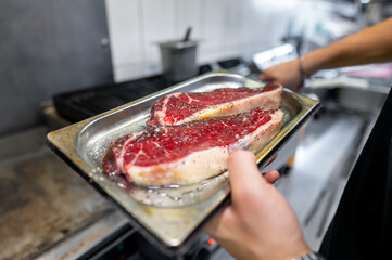 A close-up of hands holding a metal tray with a large, raw steak seasoned with salt. The focus is on the steak and its preparation, making it ideal for culinary themes