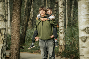 Fototapeta premium A cheerful young boy smiles as he enjoys a piggyback ride from his older brother in a serene forest setting. The tall trees in the background add a natural, peaceful atmosphere.