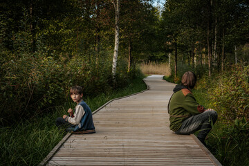 Fototapeta premium A young boy and an older teenager sit on a wooden pathway surrounded by greenery. The younger boy holds a grass blade, while the teenager looks thoughtful, gazing into the distance.