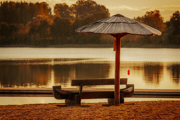 A tranquil lakeside scene at sunset, featuring a shaded bench under a beach umbrella. The golden glow reflects on the calm water, creating a peaceful atmosphere.