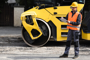 Construction worker inspecting road repair with yellow steamroller in urban area during daylight...