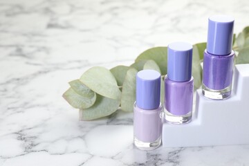 Nail polishes in bottles and eucalyptus branch on white marble table, space for text