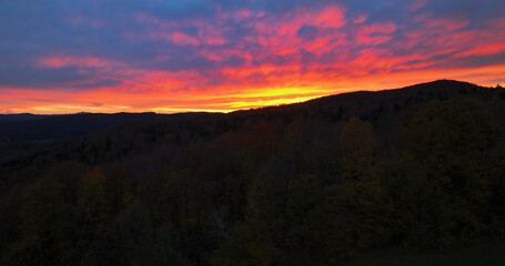 AERIAL: Spectacular aerial view of the setting sun shining on the dark countryside on an idyllic fall evening. Burnt orange autumn skies gently illuminate the forest-covered hills below the cloudy sky