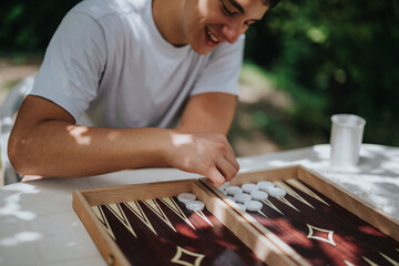 Young man plays backgammon outdoors, enjoying the strategic and social elements of the board game in a sunny, relaxing park environment.