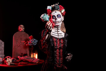 Woman posing with blue rose during Dia de los Muertos, preserving cultural traditions between generations. Terrifying lady with cyan flower wearing la Catrina costume to celebrate Mexican culture