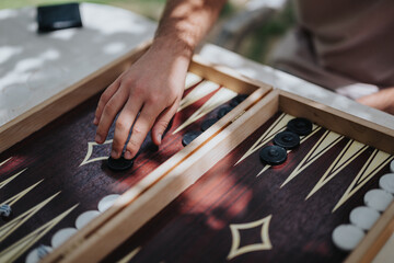 Close-up of a hand moving a piece on a backgammon board, emphasizing strategy and leisure outdoors on a sunny day.