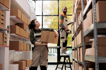 Warehouse supervisor looking at shelves full with boxes, wearing headphones listening music while carrying cardboard box in storehouse. Manager working at customers orders preparing delivery