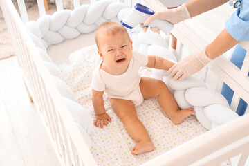 Nurse with infrared thermometer measuring crying newborn baby's temperature in crib at maternity...