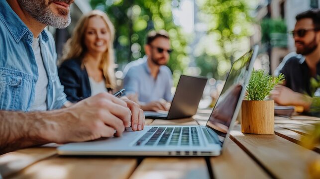 A group of professionals engage in an outdoor meeting, using laptops at a table, portraying a blend of productivity, technology, and fresh air.