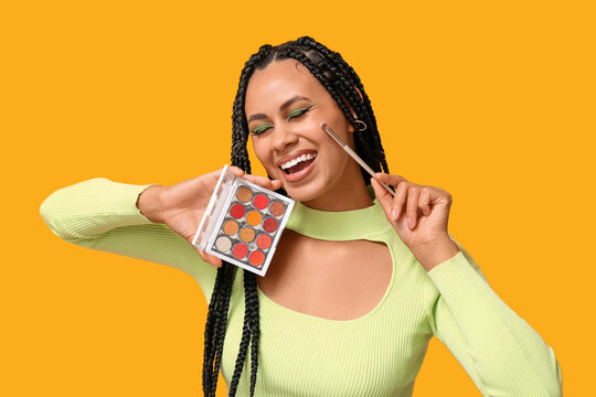 Young African-American woman with braids and beautiful makeup holding palette of eyeshadows and brush on yellow background
