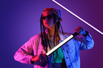 Beautiful young African-American woman with braids in sunglasses holding neon lamps on color background