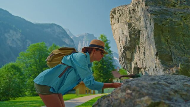 A girl tourist in a hat with a backpack drinks water from a local fountain in Switzerland, collecting it with her hands in a very beautiful picturesque place against the backdrop of mountains