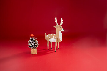 Festive holiday display on red background featuring recycling paper craft, pine cone Christmas tree plus a cardboard reindeer figurine with white spots and antlers. Eco friendly seasonal decor,