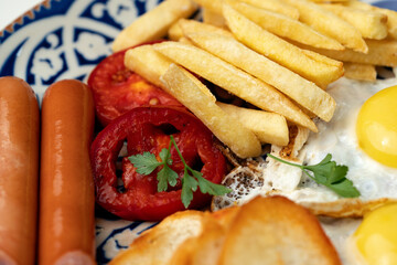 Breakfast plate with fried eggs, sausage, tomatoes, and fries served on a decorative blue plate