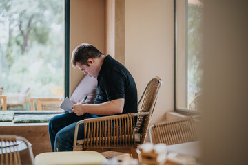 A young man with Down syndrome sits comfortably in a cozy room, intently writing on paper,...