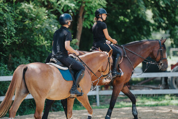 Two individuals riding horses in an outdoor equestrian arena with lush greenery in the background.