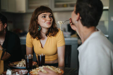 Two friends share a meal and laughter in a cozy kitchen setting, highlighting friendship and joy. A young woman smiles as her friend offers her a bite of pasta.