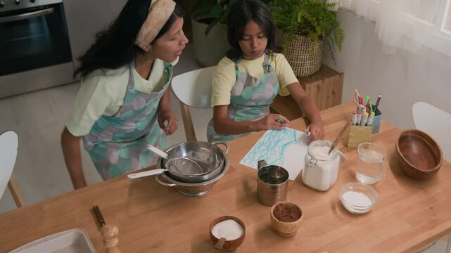 Top view shot of Latin aunt in apron and hairband bringing bowls and utensils to wooden table, preparing to make cookie dough with little niece who drawing in kitchen