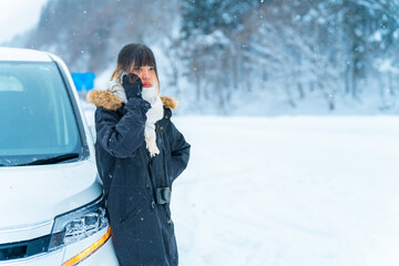 Asian woman standing outside the car using mobile phone calling assistance case of vehicle breakdown or run out of gas on the road in snow day. Car insurance, Automobile problem and garage concept.