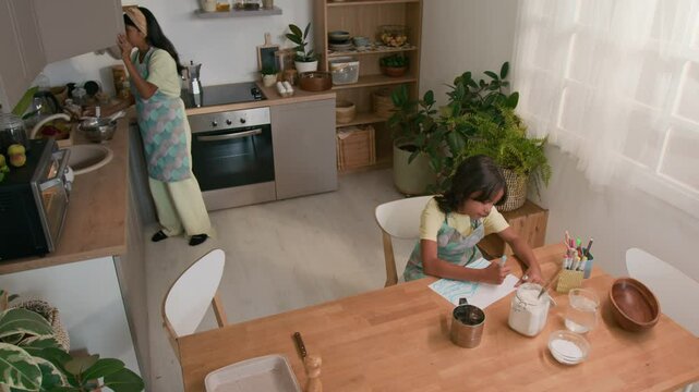 Top view of Hispanic mother in apron and hairband bringing utensils and ingredients from cupboard to table, preparing to bake cookies with little daughter who drawing in kitchen